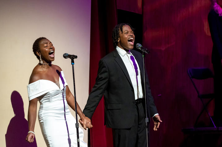 A Black female in a white dress and a Black male in a black suit singing on stage