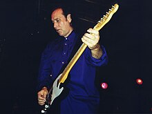 A man, wearing blue, plays an electric guitar and gazes down on the ground.