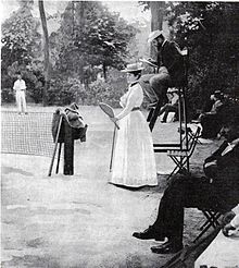 Vue d'un terrain de tennis avec une femme en robe blanche tenant une raquette et un arbitre sur sa chaise.