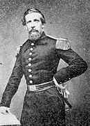 Black and white photo shows a bearded man standing with his left hand on his hip. He wears a dark military uniform with a single row of buttons and epaulettes.