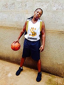 Photo d'un homme trans noir aux cheveux longs, en uniforme de basket-ball, tenant un ballon de basket dans la main.