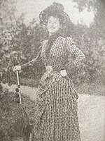 Jeune femme dans un parc avec allée et ombrelle en guise de canne, chapeau, robe longue ancienne.