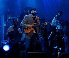 A man holding a guitar with a microphone with pedestal in front of him.