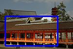 A roofed corridor and two connected buildings with gabled roofs. All of them have vermillion red wooden beams, white walls and are standing on stilts. Beyond the buildings in the background on a hill stands a five-storied red colored pagoda and a large building with gabled roof.