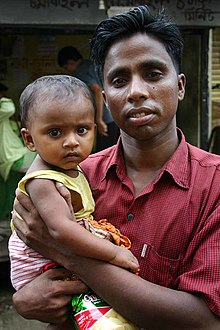 Un jeune père habillé d'une chemise en coton rose, à l'air fier mais fatigué, porte sa fille dans ses bras et sourie à l'appareil photo. La petite fille, qui porte une robe, est assise sur le bras de son père et regarde l'appareil en fronçant les sourcils.