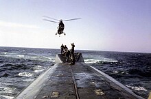 Surfaced nuclear submarine USS Triton with a six-man deck handling party standing on the raised housing located on the after end of the main deck while a U.S. Marine Corps Sikorsky UH-34 Seahorse helicopter hovering overhead.