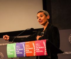 Zainab Salbi, wearing a black jacket, speaking on stage on a podium. The lectern has the logo "Girls Impact The World Film Festival"