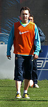 A man wearing long, dark blue shorts and a light blue sweatshirt under an orange bib.