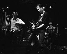 The band Flipper is performing at a club. From left to right are the singer, drummer, and electric guitarist. The singer is seated on a stool, and he is holding a pair of crutches.