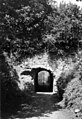 A ruined arched concrete gate in a fort wall, entered by an entrenched road