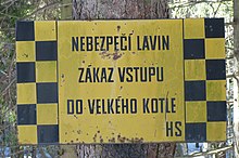 Žlutě zbarvená cedule s nápisem „Nebezpečí lavin, zákaz vstupu do velkého kotle, HS“. Levý a pravý okraj cedule jsou šachovnicově černožluté.