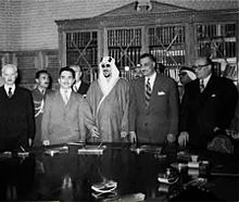 Photographie de cinq hommes debout cote à cote derrière une table. Quatre portent des costumes et le cinquième porte un costume traditionnel bédouin.