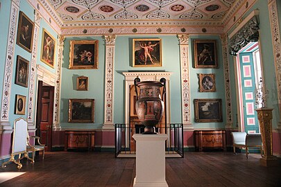 Farbfotografie eines Innenraums mit einer Gemäldegalerie an der linken und hinteren hellblauen Wand. Antike Szenen, Landschaften und Porträts sind abgebildet. In der Mitte steht auf einem weißen Podest eine dunkle Vase mit antiken Szenen. Die Wände sind mit Pfeilern versehen und haben jeweils eine Tür, die oben mit Ornamenten verziert ist. Rechts befindet sich vor dem Fenster mit blauer Seidengardine ein Chaiselongue.