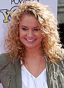 A head shot of a young woman with blonde, curly hair, wearing a white top and a khaki jacket. She is posing at a press event and smiling.