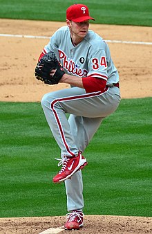 A man in a gray baseball uniform with red trim and a red baseball cap holds an unseen baseball with his right hand while wearing a black baseball glove on his left; he has one leg lifted partially in the air