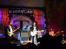 Peter, at left, plays his bass guitar and is partly turned towards Jon. Jon plays his guitar and sings into a microphone while Dann at the right is seated behind his kit.