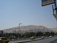 A photo a modern city with a mountain in the background. There is a road, cars, street lights, a sign, and other city infrastructure in the foreground.