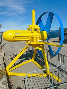 The Sabella D03 turbine. Painted bright yellow, a steel frame tripod supports the nacelle containing the generator. To the right, painted blue is the 6-bladed rotor, the tips of the blade connected by a metal band. The turbine is sat on a quayside with metal barriers around it.