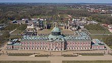 Frontale Farbfotografie in der Obersicht von einem Schloss mit vier Seitenflügeln und einer Dachkuppel. Auf den Dächern und vor dem Erdgeschoss sind Figuren aufgereiht. An den beiden äußersten Flügeln sind zwei kleine Dachkuppeln, die wie die großen Kuppelstatuen an der Spitze tragen. Im Hintergrund befindet sich ein halbrunder Säulendurchgang mit zwei Gebäuden, die Dachkuppeln und Säuleneingänge haben. Im Hintergrund sind weitere Gebäude und viel Wald mit Orten zu sehen.