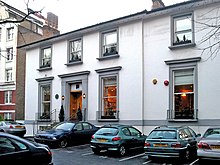 A number of cars are parked in the asphalt car-park of a two-storey white building. The building appears once to have been detached, but a two-storey extension is apparent, on the right side of the building. The ground floor contains three large sash windows, each with a small metal railing around the sill. The first floor contains four shorter sash windows. The decorative stonework around the windows and main entrance has been painted grey. Two chimney stacks are visible on the roof, at opposite ends. The staircase entrance to the building is bordered by metal railings. To the left of the image, a much larger brick building can be seen in the distance.