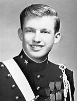 A black-and-white photograph of Trump as a teenager, smiling, wearing a dark pseudo-military uniform with various badges and a light-colored stripe crossing his right shoulder