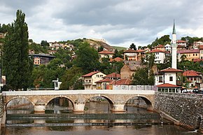 The Babića Bašča local community, with Šeher-Ćehaja Bridge in front, Inat kuća just behind it, in front of the minaret which on the other hand belongs to the Vekil-Harač mosque. Cemetery Alifakovac can be seen on the right side in the background, while the fortress on the hill in background, left on the photo, is the Bijela Tabija.