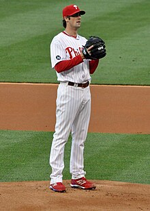 A long-haired young man in a white baseball uniform with red trim and baseball cap holds his throwing arm behind a black baseball glove.