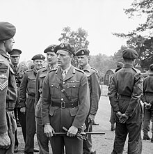 General officer with gloves and cane in hand, talking to a sergeant, surrounded by other officers and men