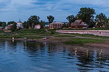 Photographie depuis le fleuve d'une berge de la ville, avec des maisons en bois et une église en arrière-plan.