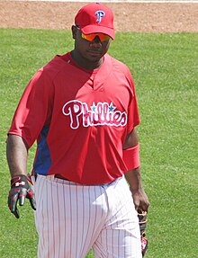 A dark-skinned man wearing a red baseball jersey with "Phillies" across the chest and a red baseball cap walking on a grass field