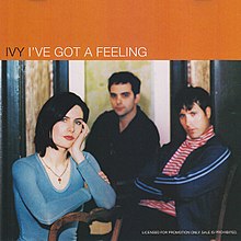 The three band members sit on chairs while the song's title and respective artist appear above them on an orange background.