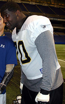 An American football player leans forward. He is not wearing pads but is wearing his white jersey over a long-sleeved gray shirt.