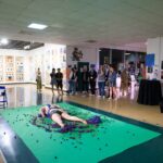 A person lays face down at CalArts on a green tarp on top of tomatoes painted purple while an audience watches.