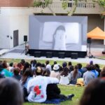 A group sits on the lawn outside, watching an animated film on a large screen.