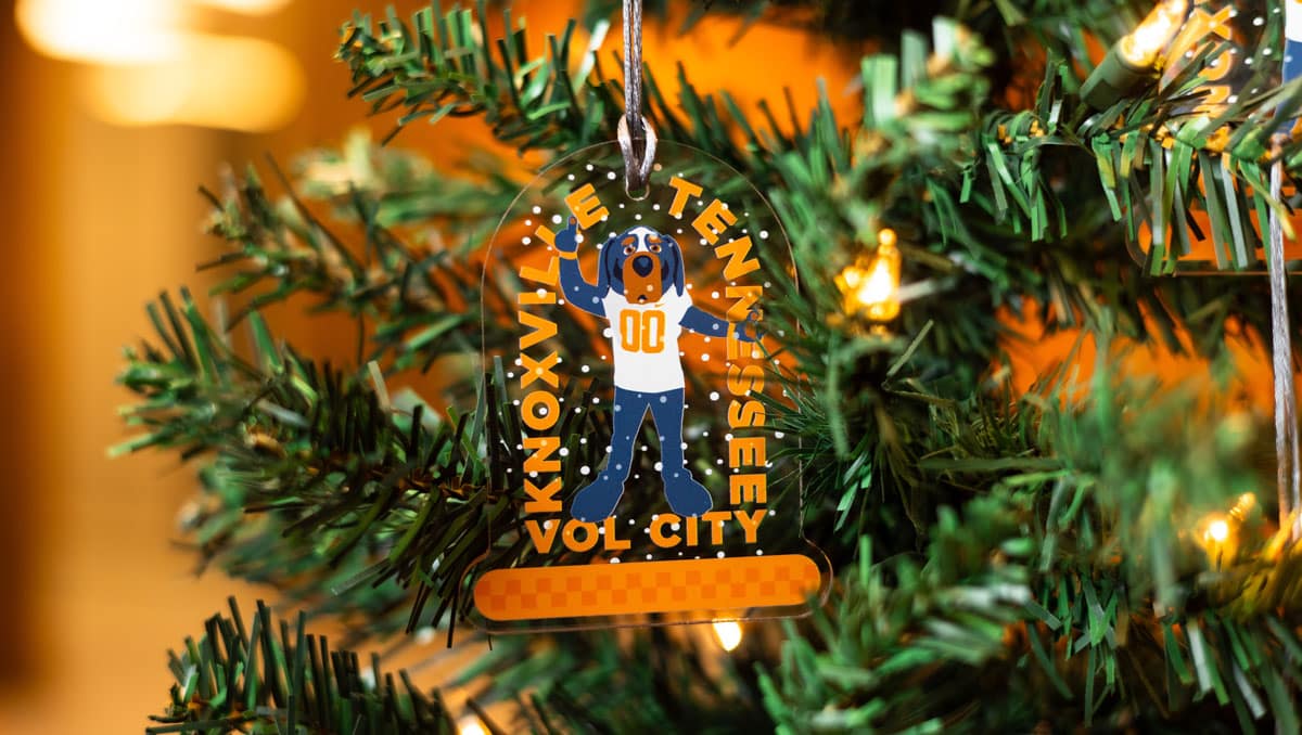 An ornament featuring Smokey surrounded by snow hangs on an evergreen tree