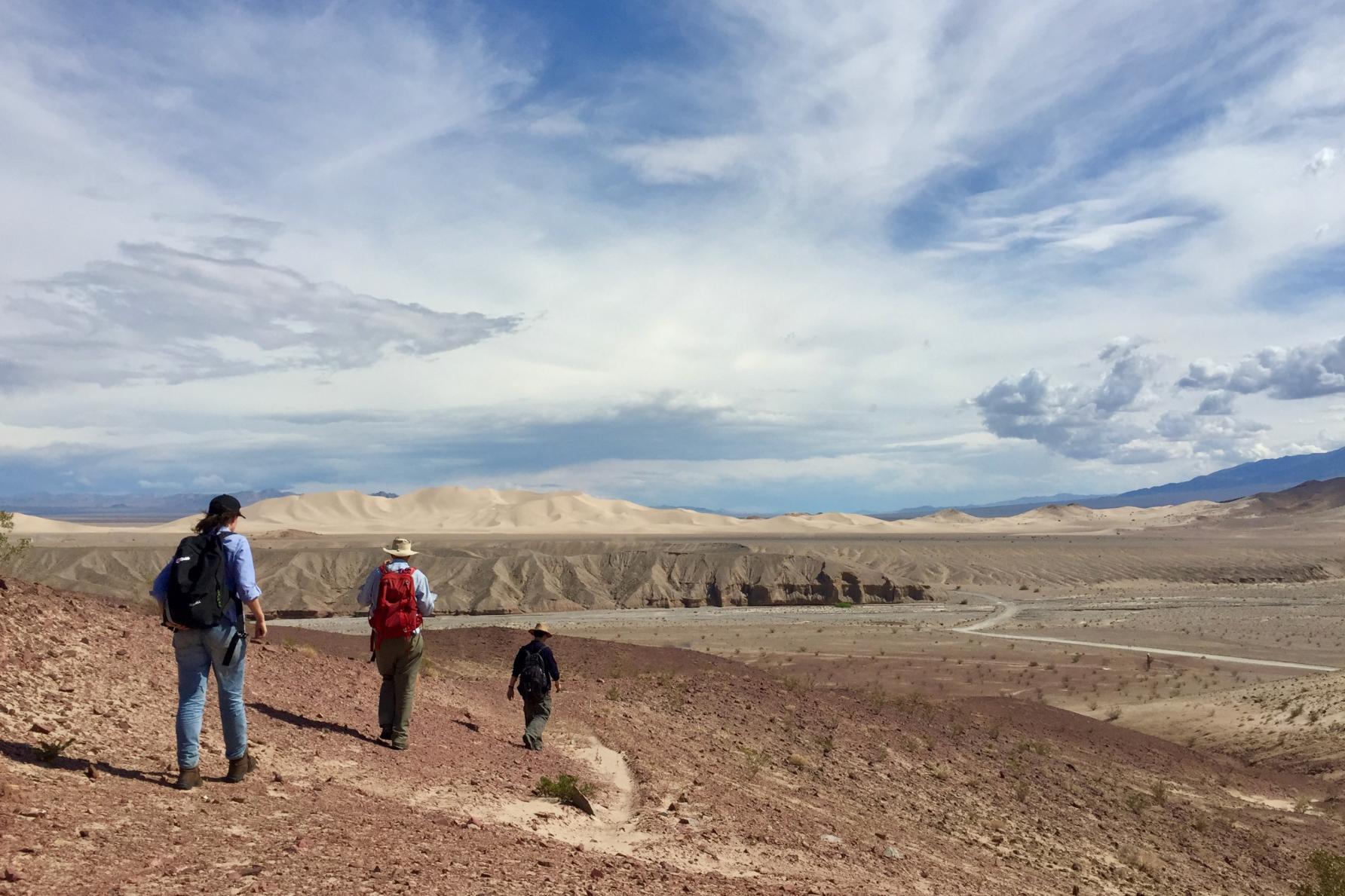 three people walk away in desert landscape under cloudy, windy sky