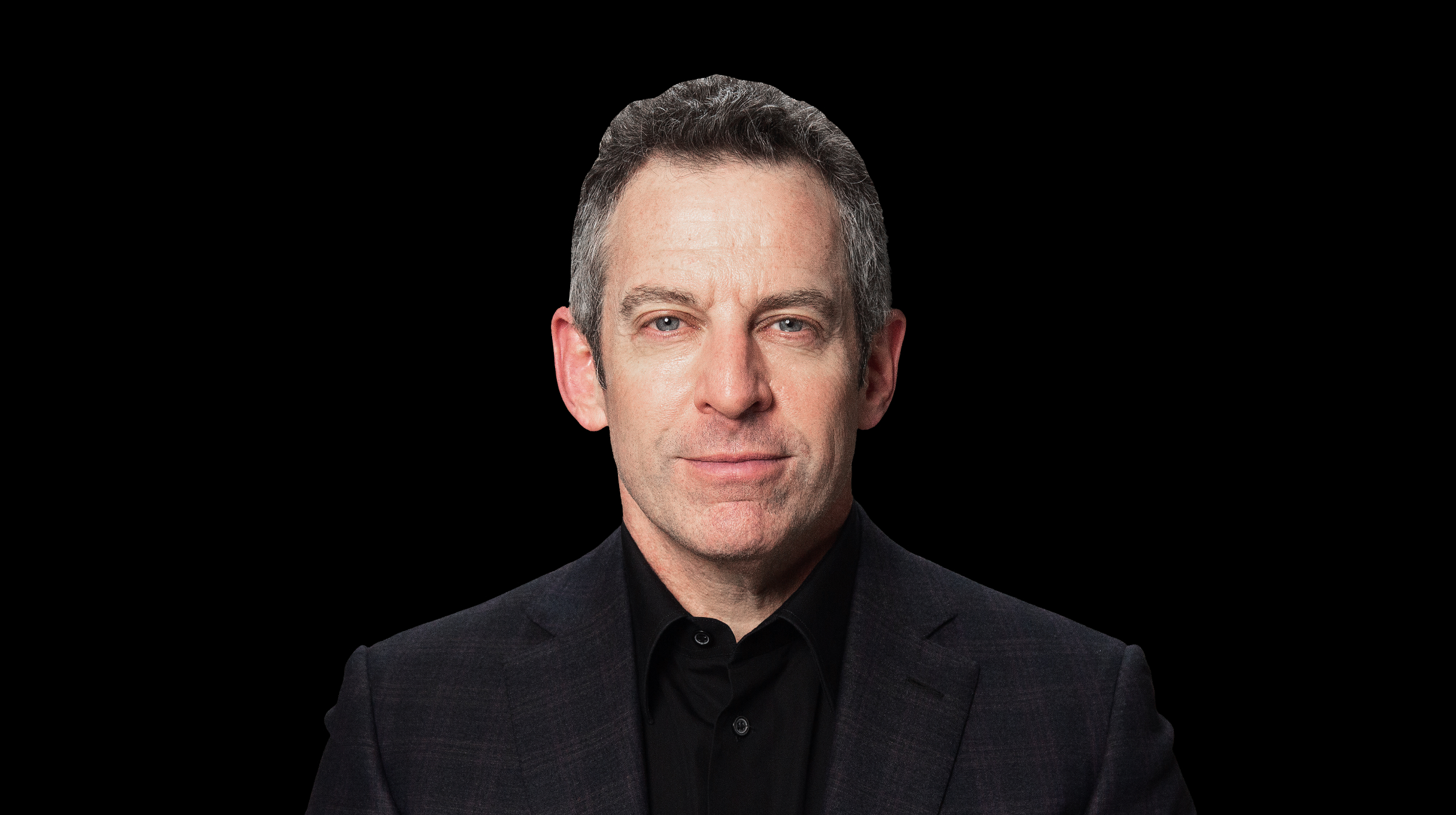 A man with short, grayish hair, wearing a dark blazer and black shirt, stares directly at the camera against a black background.