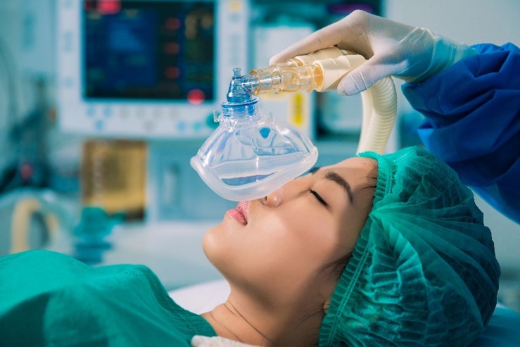 A woman wearing a hospital gown and a hair net, lying on a hospital bed as a gloved hand places a face mask emitting general anesthesia over her nose and mouth.