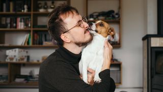 A man wearing glasses and a black roll-neck jumper is shown being licked by his dog on the face. 