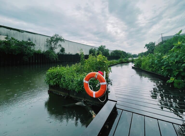 At the Wild Mile, along the Chicago River, in Chicago, facing west. A bright orange life preserver is at the center of the image, which shows the river walk on a rainy day. A white, industrial building stands in the background.