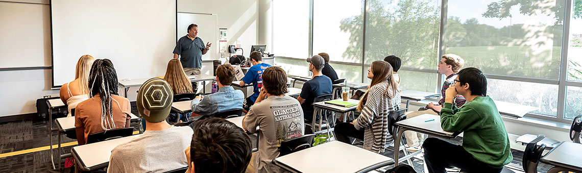 Students in class at desks listening to a teacher lecture