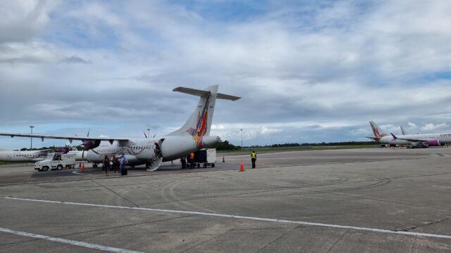 Medium-sized two-engine turboprop aircraft on tarmac runway, facing away from the photographer, under slightly cloudy blue skies. Caribbean Airlines livery (colourful hummingbird on tail).