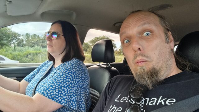 Ruth drives a car; Dan, in the passenger seat, looks into the camera as he takes a selfie.