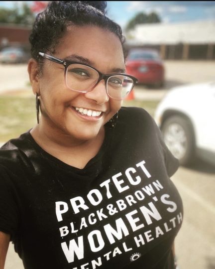 Snapshot of Irene in glasses smiling and wearing a black t-shirt with white lettering 'Protect black & brown women's mental health'. 