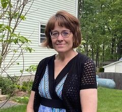 Tina has light skin tone, short brown hair, and glasses. She is wearing a blue dress and standing outside with green grass behind her. 