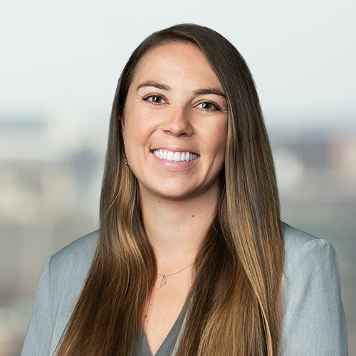 Professional headshot of Kayla, a woman with light skin tone and long brown hair. Kayla is smiling and wearing a gray suit jacket.