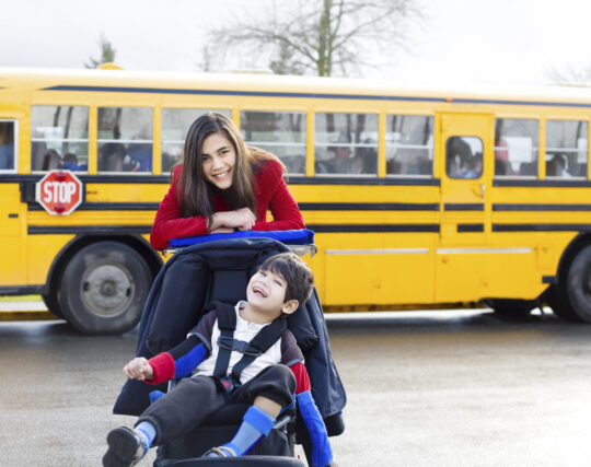 Young child with light skin tone smiles happily while young adult pushes them in an adapted stroller. A yellow school bus is visible in the background. 