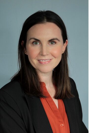 Megan smiles at the camera in a professional headshot. She has light skin tone, dark hair, and is wearing rust colored shirt and black suitcoat.