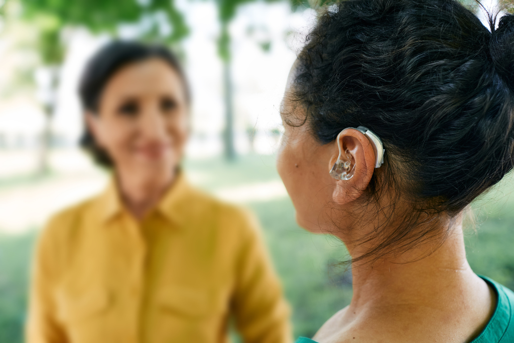 Two people with medium skin tone face each other. The person closer to the viewer is facing away from the camera and their dark hair is pulled up and their hearing aid is visible behind their ear. The other person is facing the camera but is out of focus but their smile is as bright as their yellow shirt.