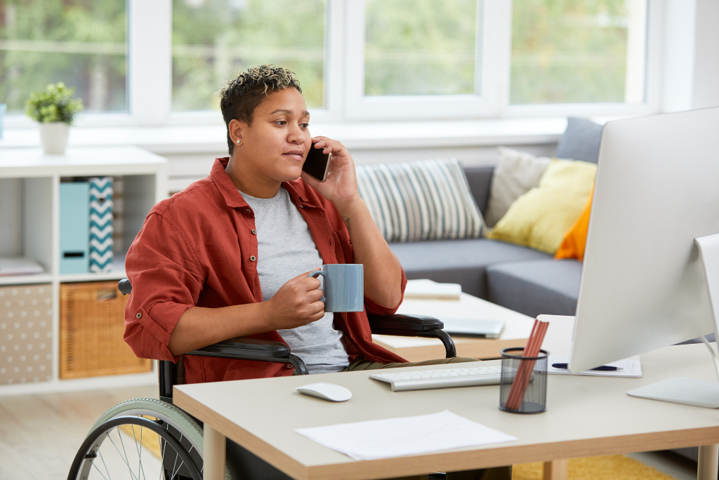 Person with medium skin tone and short hair talks on their cell phone while sitting in their wheelchair at their desk from a modern and well lit home office.